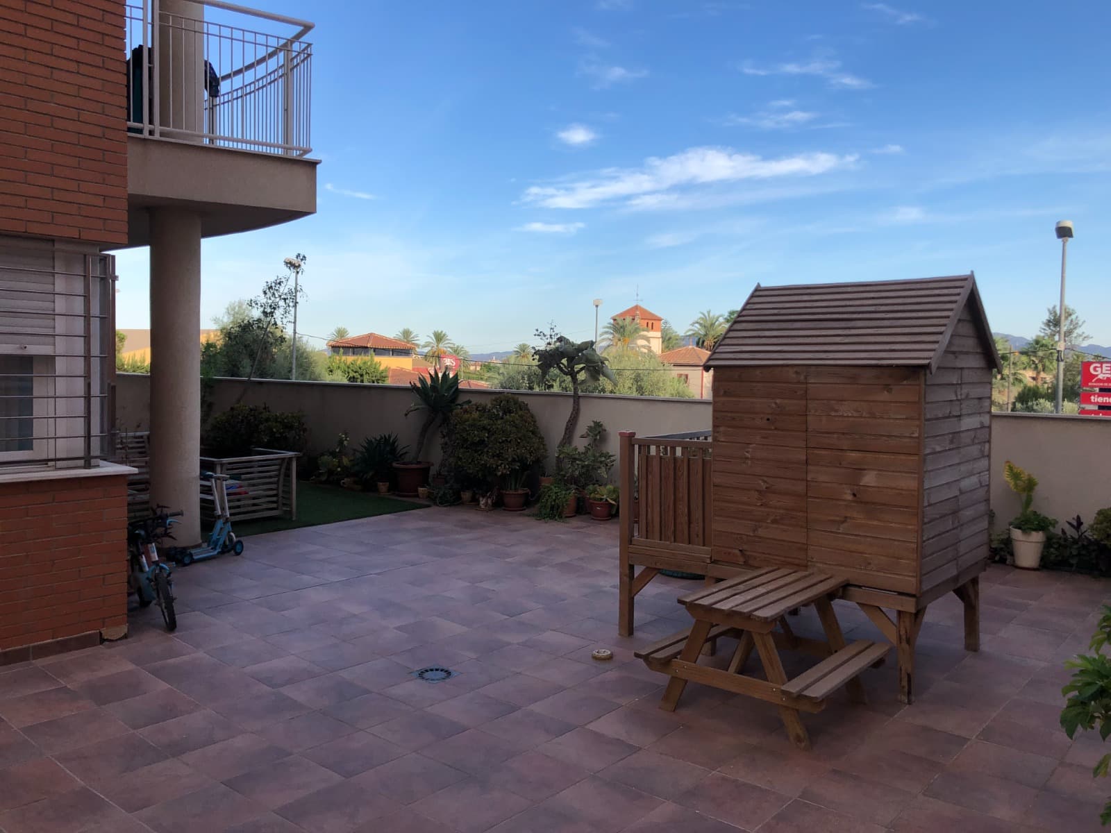 Patio exterior pavimentado con casita de madera infantil, mesa de picnic y zona ajardinada